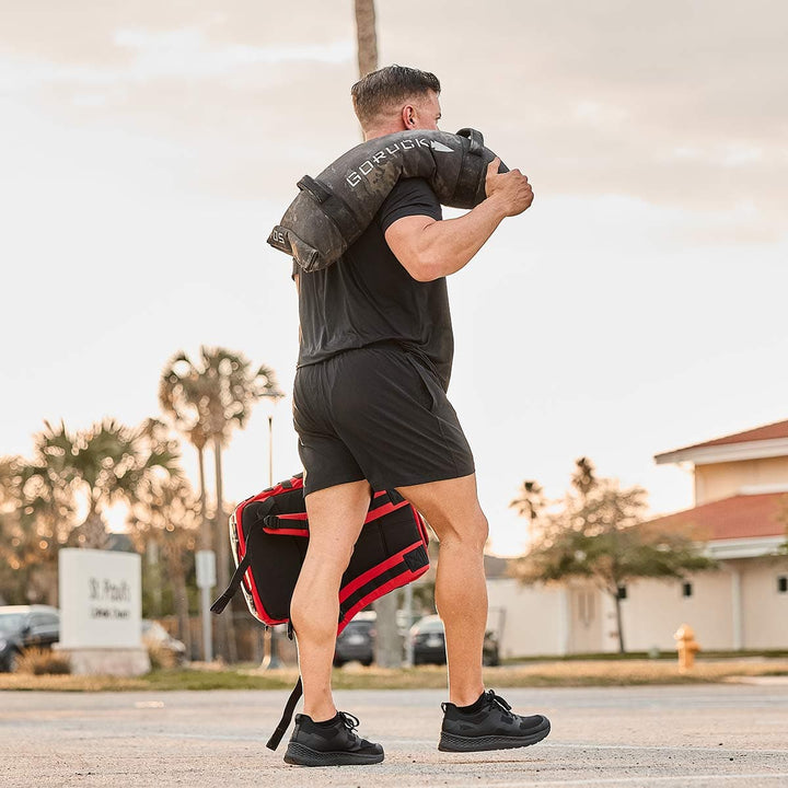 Clad in GORUCK's Men’s Training Shorts - ToughStretch, a person carries a heavy sandbag on their shoulders while walking outdoors. The setting sun casts a warm glow over the palm trees and buildings in the background, complementing their black athletic wear.