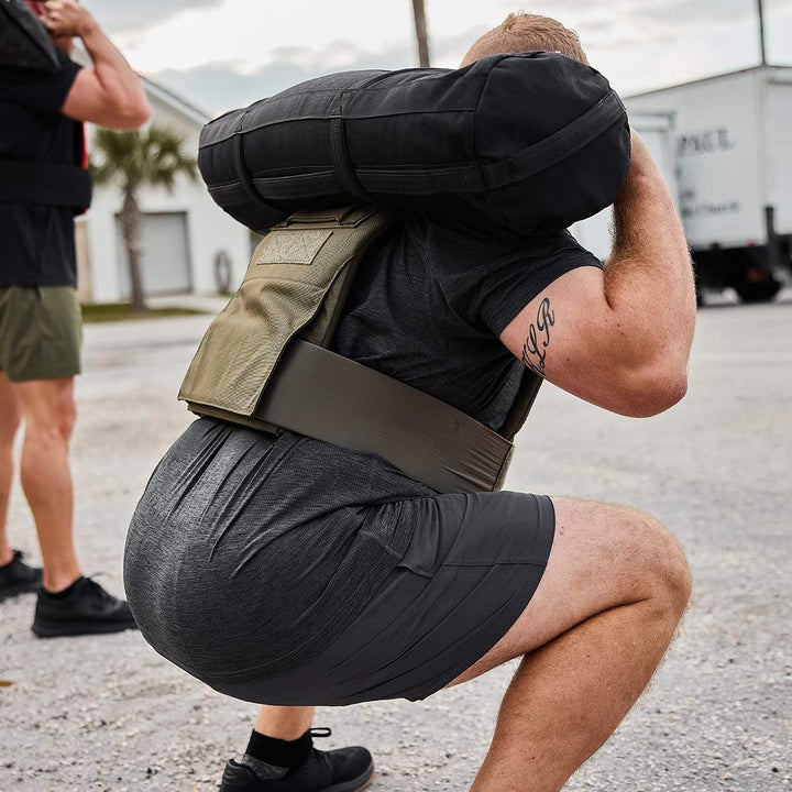 A person squats outdoors, lifting a heavy sandbag onto their shoulders while wearing GORUCK's Men’s Training Shorts - ToughStretch and a weight vest, alongside another individual in similar gear. The scene takes place in a parking lot with a truck nearby, showcasing the durability promised by our SCARS Lifetime Guarantee.