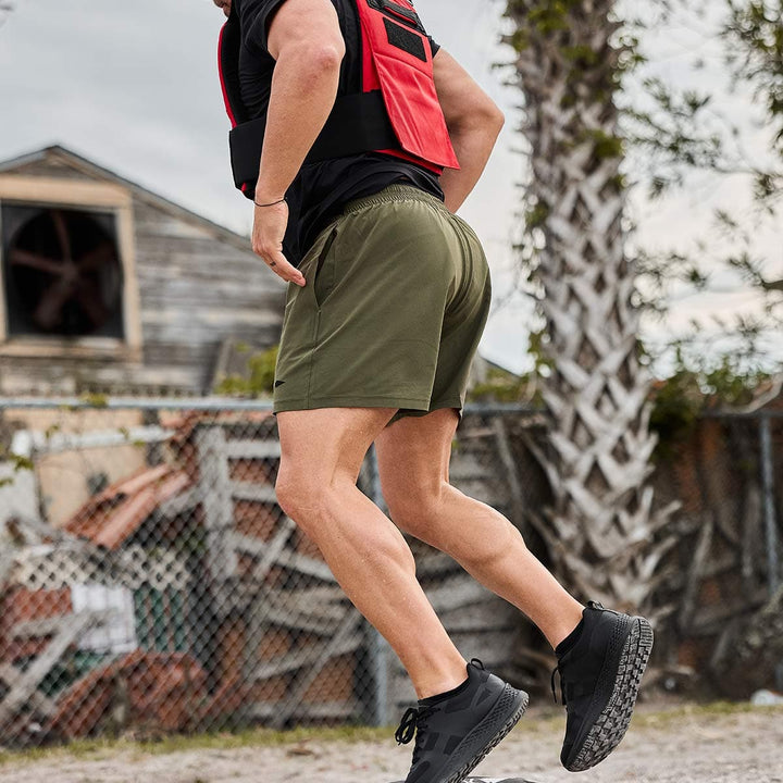 A person running outdoors is wearing a red and black vest, Men’s Training Shorts in green by GORUCK, made with ToughStretch fabric for optimal comfort, and black sneakers. They are on a dirt path with a rustic wooden building and a palm tree in the background.