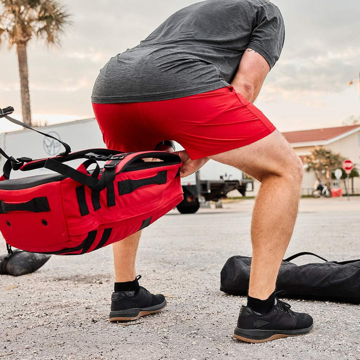 Clad in a gray shirt and GORUCK's Men’s Training Shorts made of ToughStretch fabric, an individual bends down to retrieve a red duffel bag. In the background, trees, buildings, and vehicles fill the scene of the paved outdoor space.