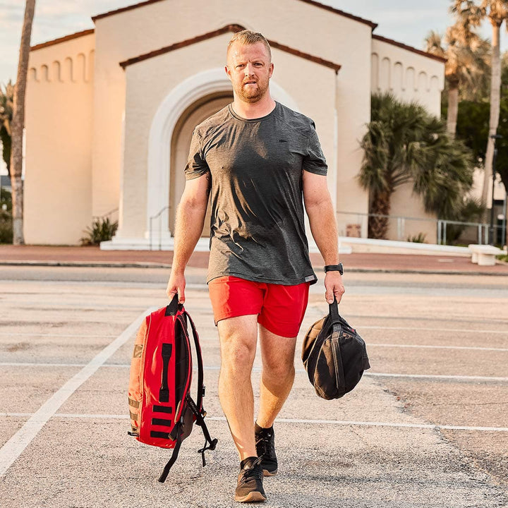 A man strolls through a parking lot, carrying two bags, one in each hand. He sports a gray T-shirt, GORUCK Men’s Training Shorts - ToughStretch, and black sneakers. Behind him stands a building with an arched entrance framed by palm trees. The sky is partly cloudy, suggesting a sunny day to come.