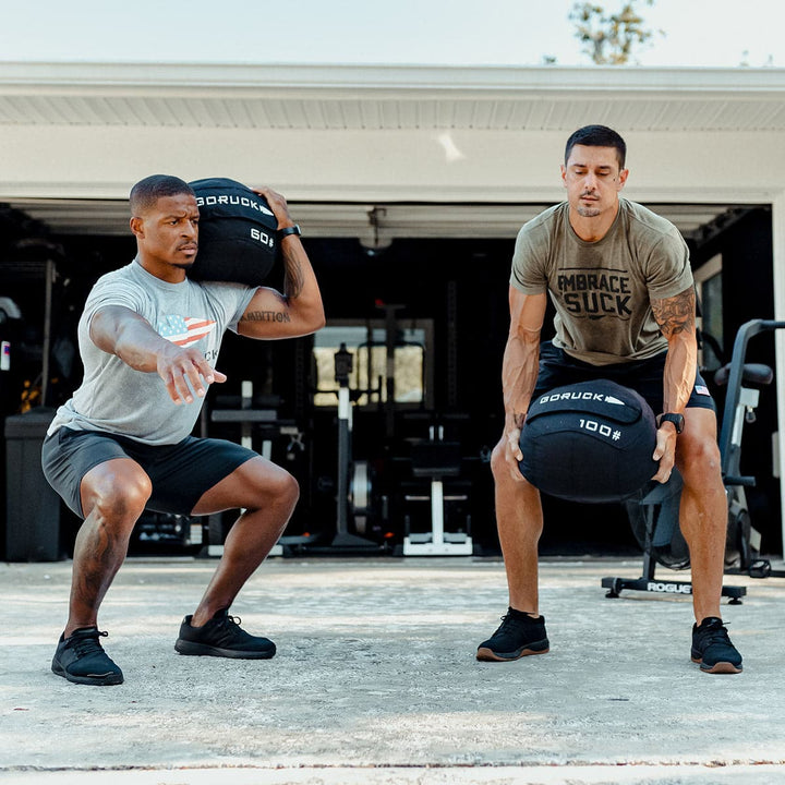 Two men energetically improving their core stability with GORUCK Sand Medicine Balls, working out in front of a garage.