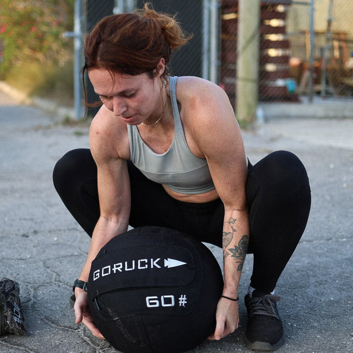 A woman in athletic wear is outdoors lifting a 60-pound GORUCK Sand Medicine Ball, boosting her core stability.