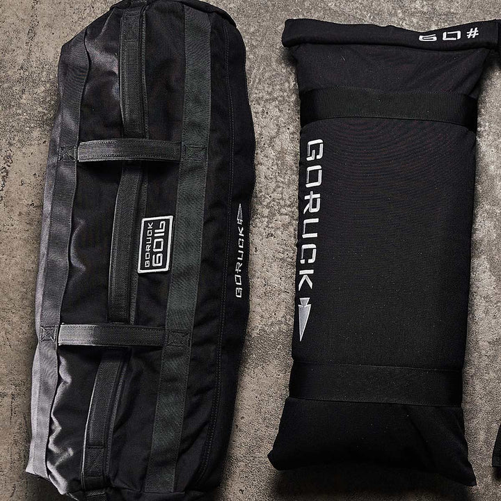 Two black GORUCK Sandbag Kits with Filler Bags sit on concrete, featuring heavy-duty handles and the Scars Lifetime Guarantee promising durability.