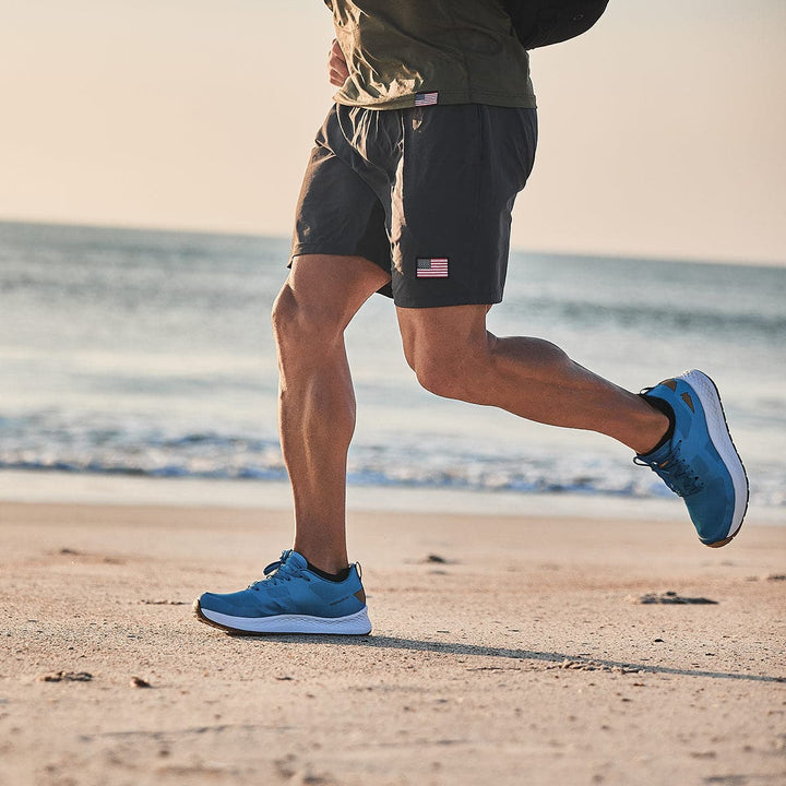 A person adorned in athletic gear sprints along a sandy beach at sunset, capturing the essence of a GORUCK Rough Runner. They sport a gray shirt, black shorts featuring a small flag emblem, and the Tidal Blue Men's Rough Runner shoes by GORUCK, crafted with Gradient Density EVA for superior comfort. The ocean expands under the gentle, warm sky.