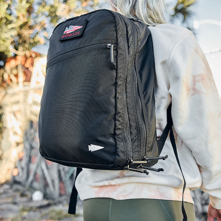 A person carrying the GORUCK Bullet Ruck Double Compartment travel backpack, made from sleek ripstop nylon, strolls outdoors near a fence and trees.