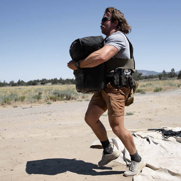 A man in sunglasses and a tactical vest performs loaded carries, hauling a GORUCK Sand Tombstone across a sandy outdoor area under a clear blue sky. He wears a gray shirt, brown shorts, and sneakers. Trees and hills are visible in the distant background.