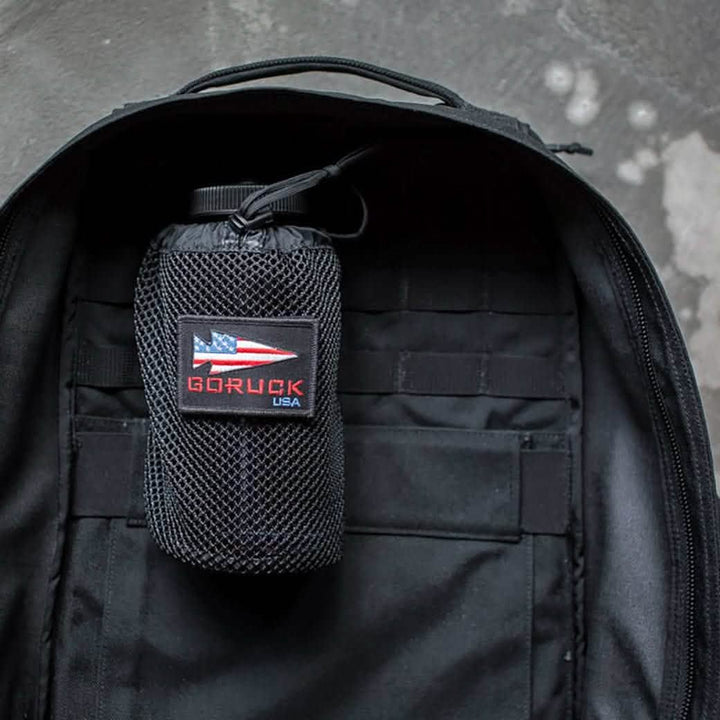 A black backpack is open with a GORUCK Mesh Water Bottle Pocket attached inside, perfect for on-the-go grab-ability. The pocket features a patch of an American flag and the text GORUCK USA. A Nalgene water bottle fits snugly in the side compartment. The background is a concrete surface.