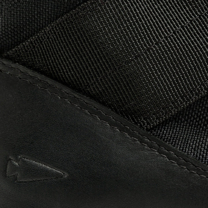 Close-up of black fabric and leather featuring a stitched arrow-shaped logo, highlighting the texture and material details, reminiscent of the rugged design characteristic of GORUCK's MACV-2 - Mid Top - Black.