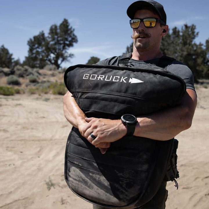 A person wearing sunglasses and a cap navigates the sandy landscape, effortlessly handling a GORUCK Sand Tombstones bag. Dressed in a gray t-shirt, they showcase their strength with loaded carries, while a watch glints on their wrist amidst scattered trees.