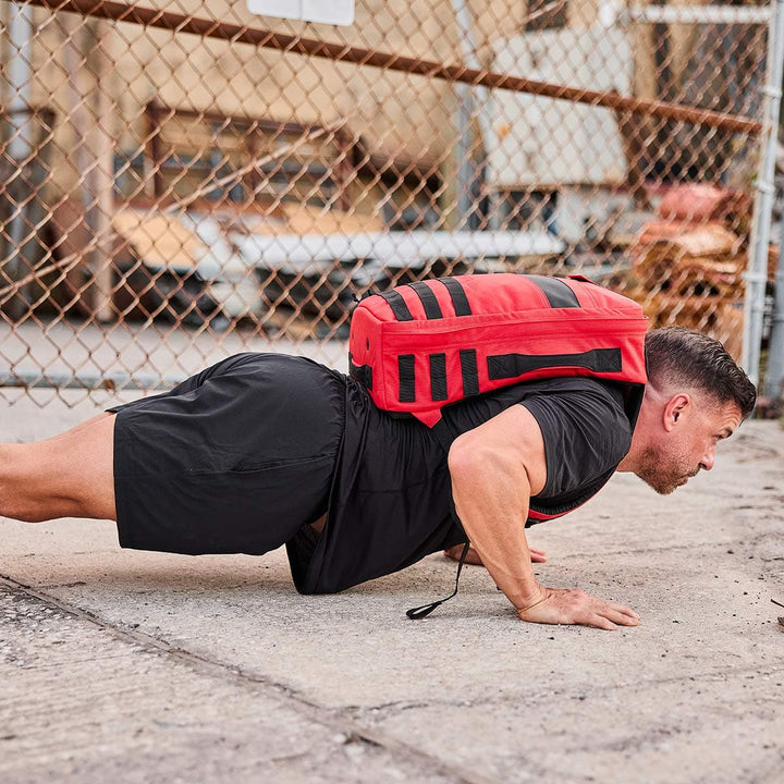 A man, sporting GORUCK’s Men’s Training Shorts - ToughStretch, performs a push-up on the concrete outdoors near a chain-link fence. His shorts are designed with durable ToughStretch fabric and come with the SCARS Lifetime Guarantee. He also carries a red backpack.