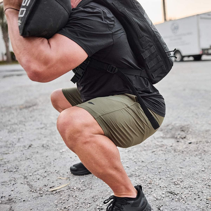 A person wearing a black shirt and olive GORUCK Men’s Training Shorts - ToughStretch fabric is squatting outdoors with a heavy backpack. The ground appears gravelly, and vehicles are visible in the background, highlighting the durability required for an adventure supported by SCARS Lifetime Guarantee.