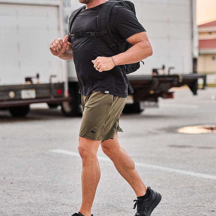 A jogger dressed in GORUCK's ToughStretch fabric training shorts, wearing a black t-shirt and shoes, carries a black backpack. Parked trucks in the background enhance the scene's dynamic energy.