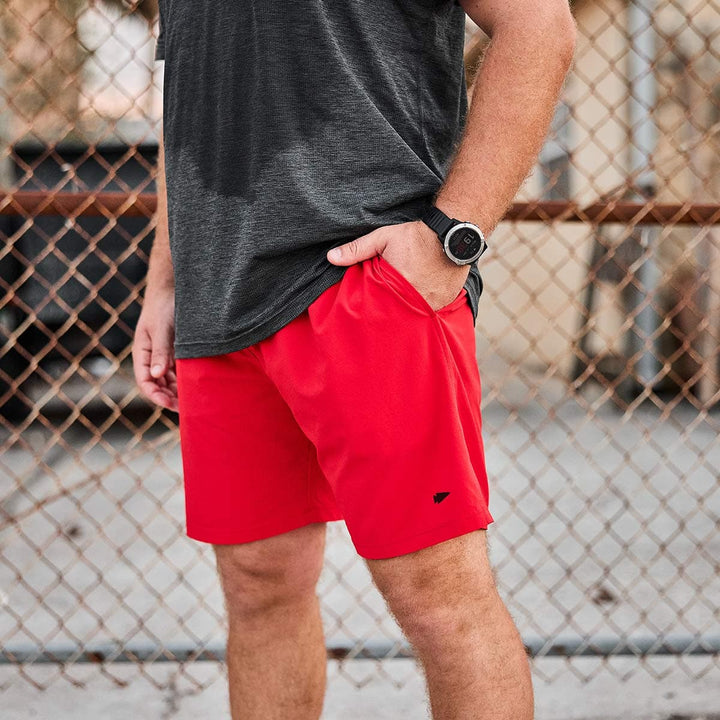 A person wearing a gray shirt, Men’s Training Shorts - ToughStretch in red by GORUCK, and a black wristwatch stands in front of a chain-link fence. Their left hand is in their pocket, showcasing the reliability of SCARS Lifetime Guarantee.