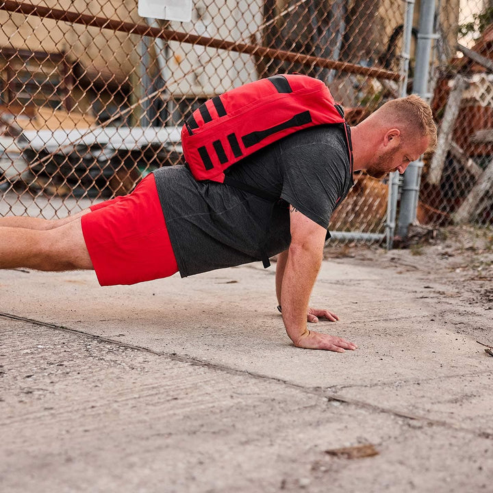 A man executes a push-up on a concrete surface, highlighting his GORUCK Men’s Training Shorts - ToughStretch. He carries a red backpack and wears red shorts along with a dark t-shirt. In the background, a chain-link fence and industrial equipment emphasize the durability associated with the SCARS Lifetime Guarantee.