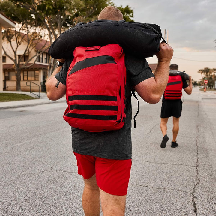 Two people walk down a street carrying sandbags on their shoulders. The person in the foreground moves with ease wearing Men’s Training Shorts - ToughStretch from GORUCK and a red backpack. The sky is overcast, and houses line the street, creating an atmosphere of determination and endurance.