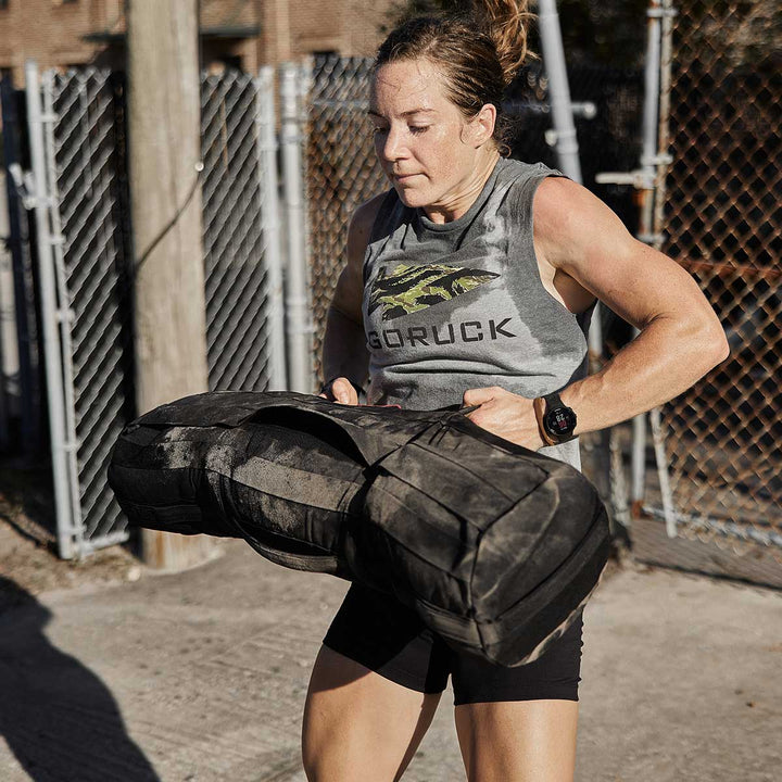 A person lifting a hefty Sandbags 2.0 by GORUCK outdoors, demonstrating their strength in a sleeveless shirt and shorts. The sandbag is equipped with neoprene padded handles for comfort, set against a sturdy fence backdrop, and every workout backed by the Scars Lifetime Guarantee.