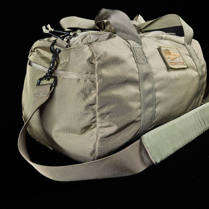 Introducing the GORUCK Gym Bag - Ripstop ROBIC®: A tan bag designed with a shoulder strap and adorned with a subtle logo patch on a black background, meticulously crafted from durable ROBIC Ripstop Nylon and featuring an integrated shoe compartment for enhanced convenience.