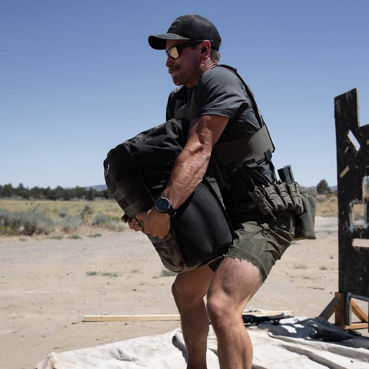 Under the clear blue sky, a man in tactical gear, sunglasses, and cap lifts a GORUCK Sand Tombstone. Clad in a gray t-shirt and green shorts, he embraces the challenge of loaded carries on the sandy terrain.