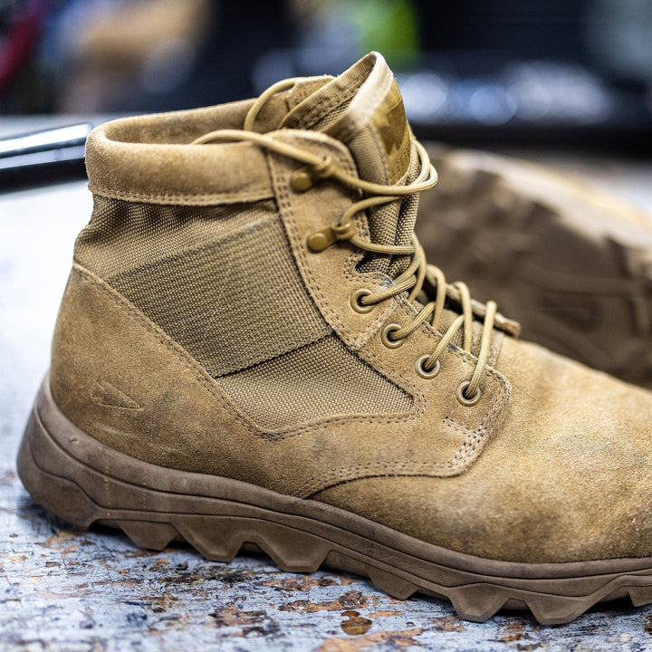 A close-up of a tan, rugged MACV-2 - Mid Top - Coyote leather boot by GORUCK, featuring thick laces and a triple compound outsole, resting on a wooden surface. A second identical boot is partially visible in the background, slightly out of focus.