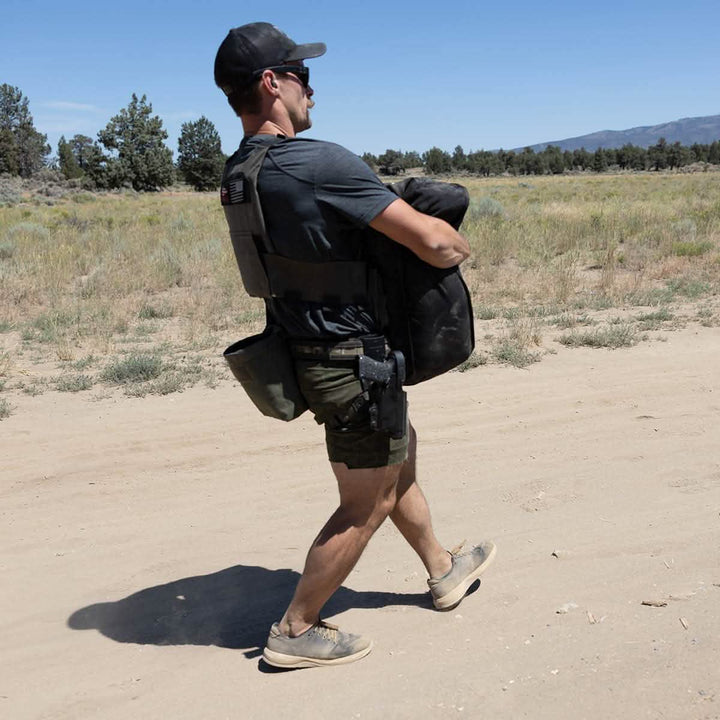 A man sporting a cap, sunglasses, and a tactical vest strides across a sandy landscape with sparse vegetation, carrying the GORUCK Sand Tombstones—a heavy black bag equipped with a multilayer closure system. Wearing a gray shirt, green shorts, and sneakers, the clear sky overhead accentuates his endurance as he maneuvers loaded carries over the flat terrain.