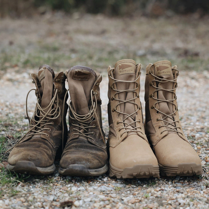 Two pairs of boots rest side by side on a gravel path. The pair on the left is worn and scuffed, while the pair on the right looks like GORUCK's MACV-2 - High Top in Coyote, featuring a triple compound outsole and appearing new and clean. Both are in a similar tan color, with grass visible in the background.