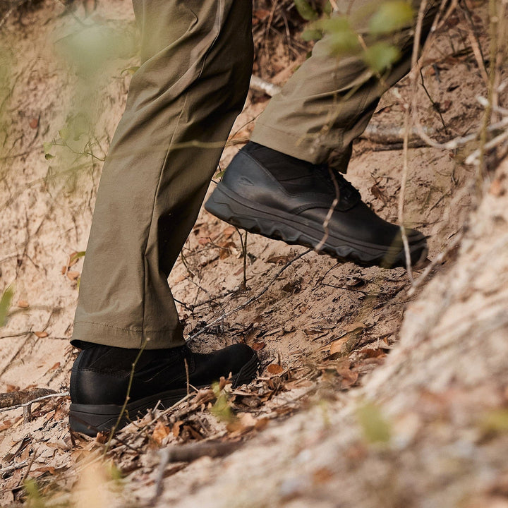 A close-up shows someone's legs in green pants, sporting GORUCK's MACV-2 Mid Top boots in black, featuring a triple compound outsole. They're navigating a steep, leaf-covered dirt path surrounded by sparse greenery in a natural outdoor environment.