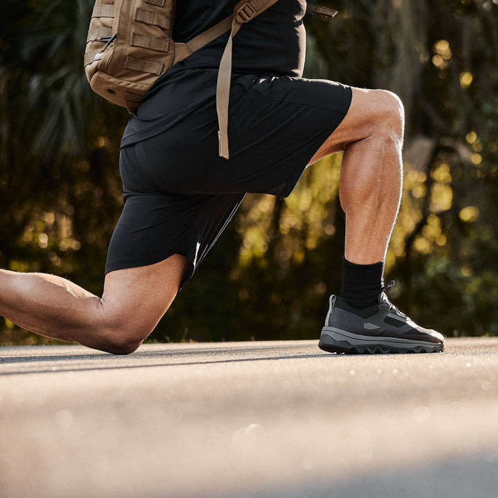 A person wearing GORUCK's Men’s Training Shorts made from ToughStretch fabric is kneeling on a paved path. With their brown backpack beside them, sunlight filters through the trees in the background, while their shoes with SCARS Lifetime Guarantee ensure lasting quality.