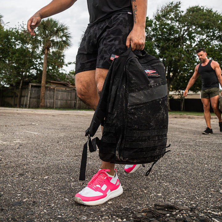 Two people are rucking outdoors; one carries a muddy Rucker 4.0 by GORUCK and wears pink sneakers.