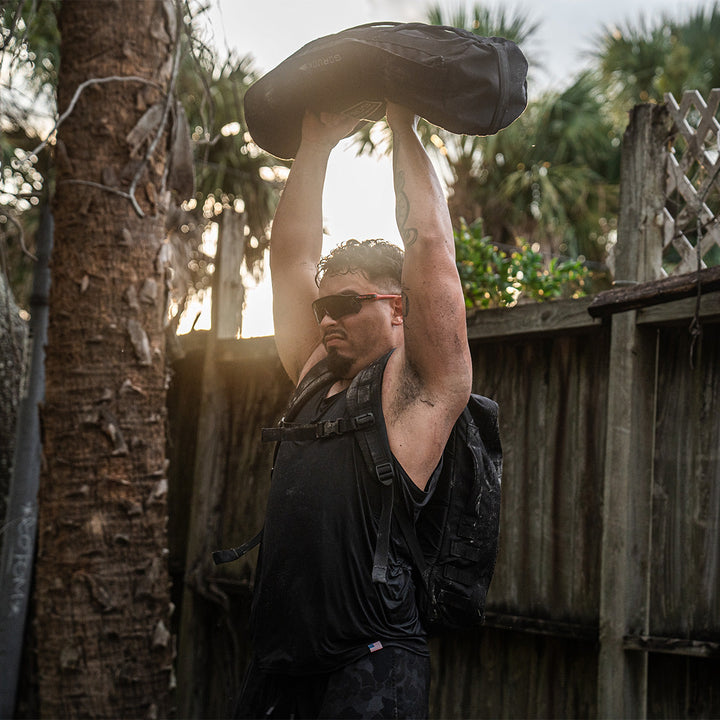 A person lifting a heavy bag overhead, wearing a GORUCK Rucker 4.0 backpack, stands in an outdoor setting with trees and a wooden fence.