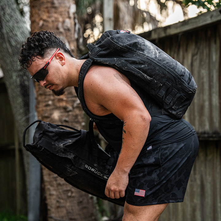 Sporting the GORUCK Rucker 4.0 while holding two black sandbags outdoors, a man wearing sunglasses and a weighted vest perfectly embodies the spirit of rucking.