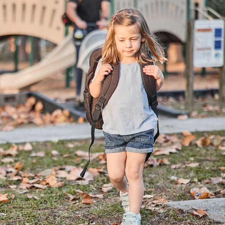 A young girl strolls through the park, her GORUCK KR1 2.0 - Kid Ruck, featuring reinforced stitching, bouncing lightly with each step as the playground looms invitingly in the background.