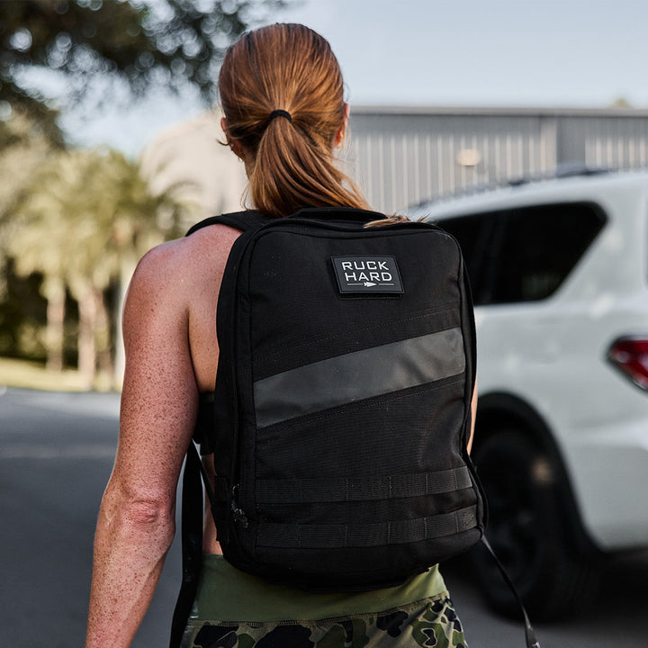 A person in a tank top, carrying a GORUCK Rucker 4.0 rucksack, is walking near a white vehicle and palm trees.