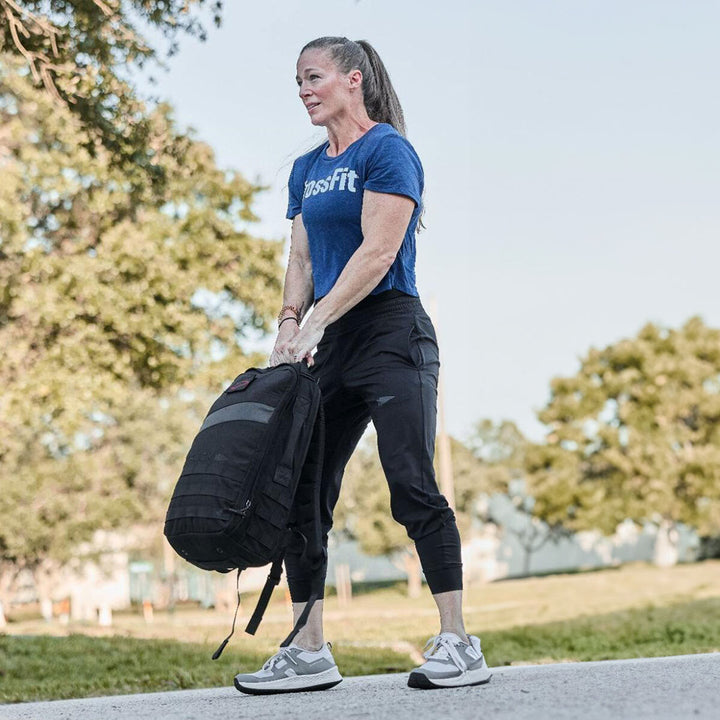 A person in a blue shirt is lifting a large black Rucker 4.0 by GORUCK outdoors on a sunny day, embracing the art of rucking.