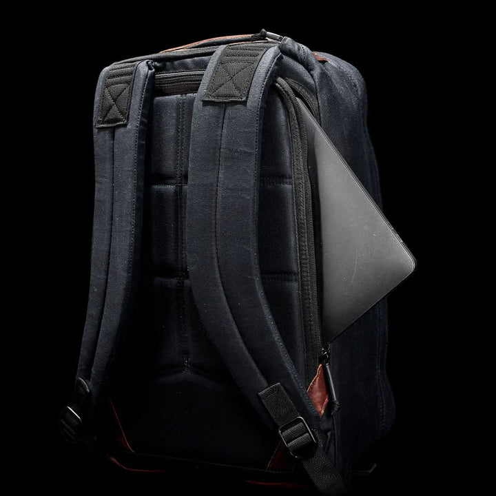 The GR1 USA - Heritage by GORUCK is a black rucksack with padded straps, made from durable waxed canvas, featuring a partially open pocket displaying a laptop against a black background.