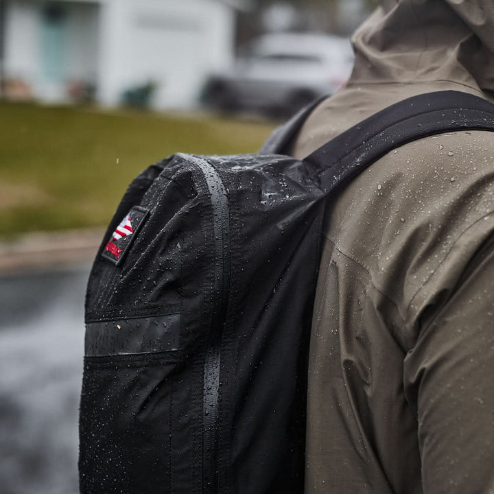 A person in a raincoat braves the rain, carrying a waterlogged Packable Bullet Ruck made from durable Ripstop ROBIC® nylon.