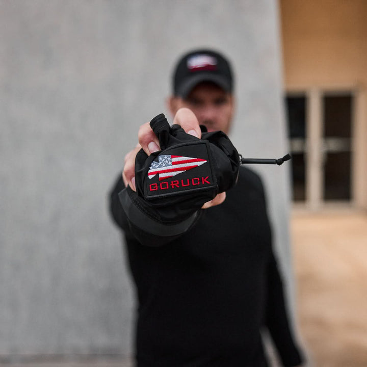 Someone holding a Packable Bullet Ruck - Ripstop ROBIC® faces the camera, dressed in a sleek black outfit and cap, with a blurred backdrop.