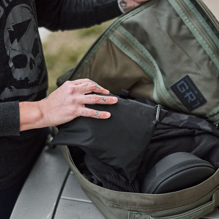 A person is carefully packing a black pouch into the Packable Bullet Ruck - Ripstop ROBIC® green backpack, which is open on the table.