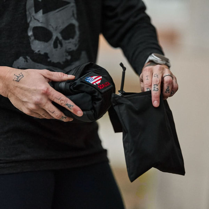 With tattooed fingers, the person carefully packs a boxing glove into a sleek black Packable Bullet Ruck made from durable Ripstop ROBIC® nylon.