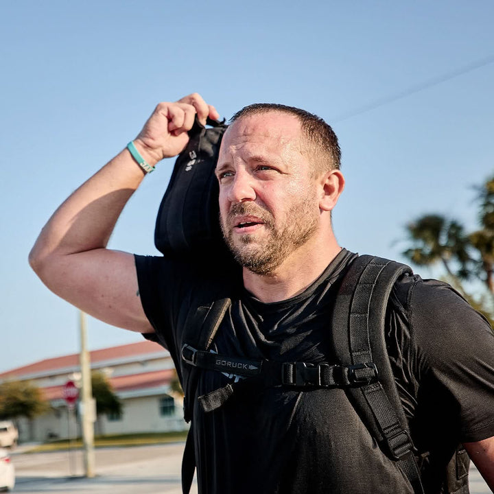 A man wearing a GORUCK Men’s Performance Tee - ToughMesh and carrying a backpack strolls outdoors, with another bag slung over his shoulder. The backdrop of a clear blue sky, trees, and buildings perfectly highlights the quick-drying performance fabric of his attire.