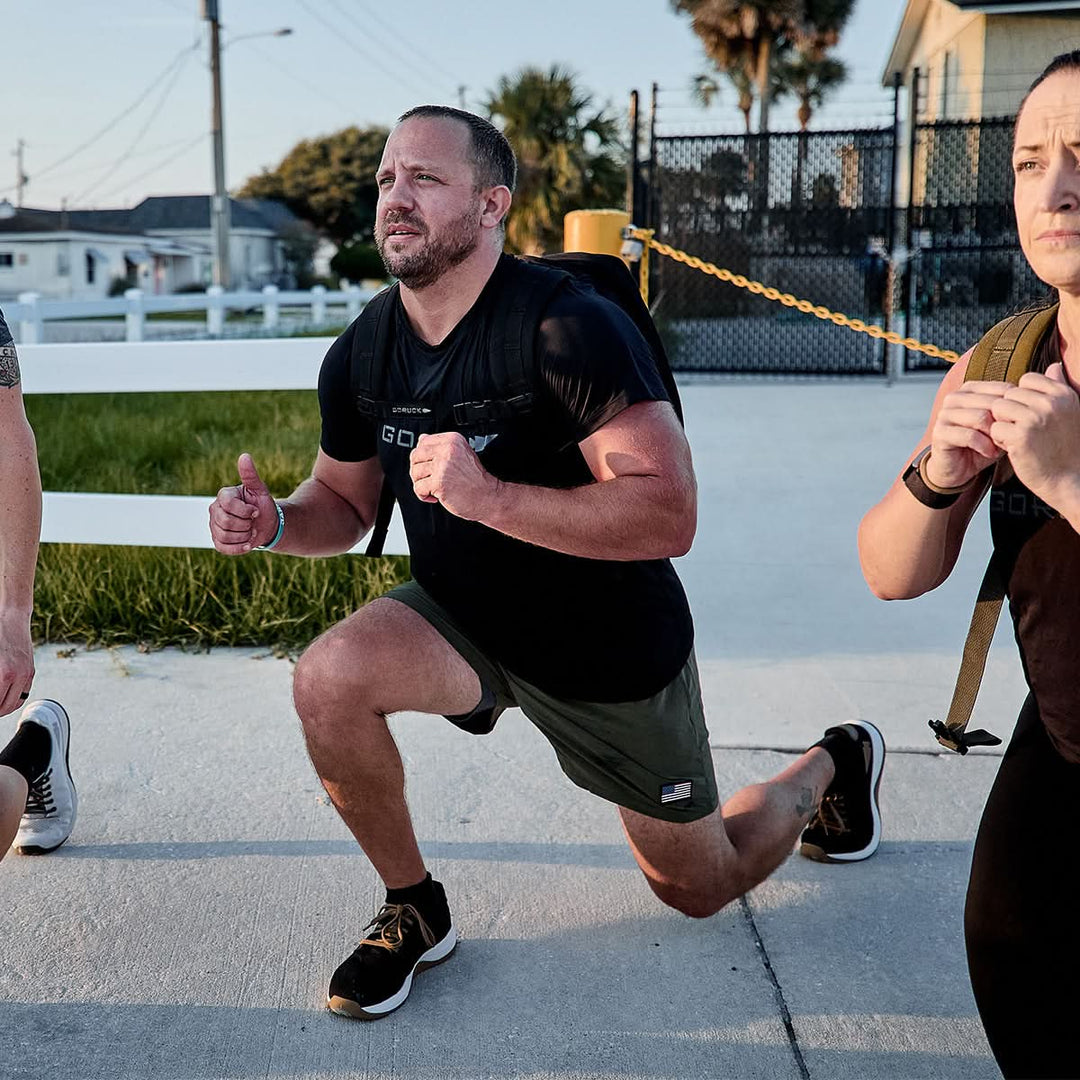 Three individuals exercise outdoors, performing lunges while wearing weighted backpacks and sporting the Men’s Performance Tee - ToughMesh by GORUCK. They are on a paved area with grass and buildings in the background, enjoying the sunny weather.