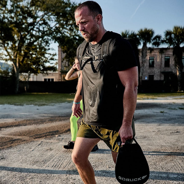 A man wearing a black Men’s Performance Tee - ToughMesh by GORUCK and green shorts carries a weight bag with the GORUCK label in one hand as he walks outside. A woman in vibrant green pants follows in the background. Trees and buildings are visible beneath a clear sky, highlighting an active day.