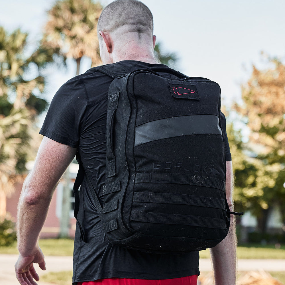 A person with a shaved head, carrying a black Rucker 4.0 by GORUCK and wearing red shorts, strolls outdoors with trees in the background.