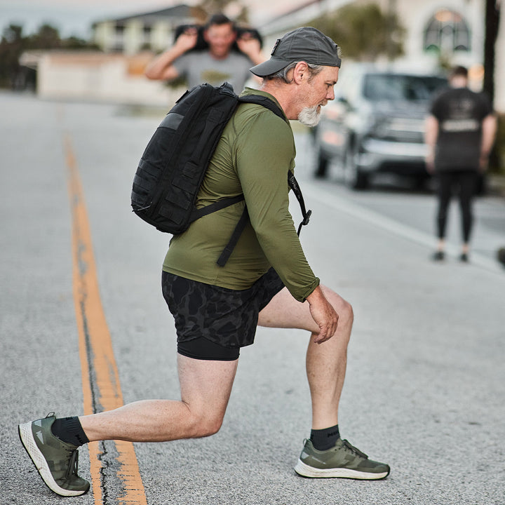 A man in exercise gear performs lunges on a bustling street, wearing a GORUCK Rucker 4.0 backpack and cap; others participate in rucking exercises in the background.