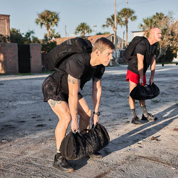 Amidst gently swaying palm trees, two people perform a sandbag exercise outdoors. Nearby, a GORUCK Rucker 4.0 rucksack awaits a challenging day of post-workout rucking adventures.