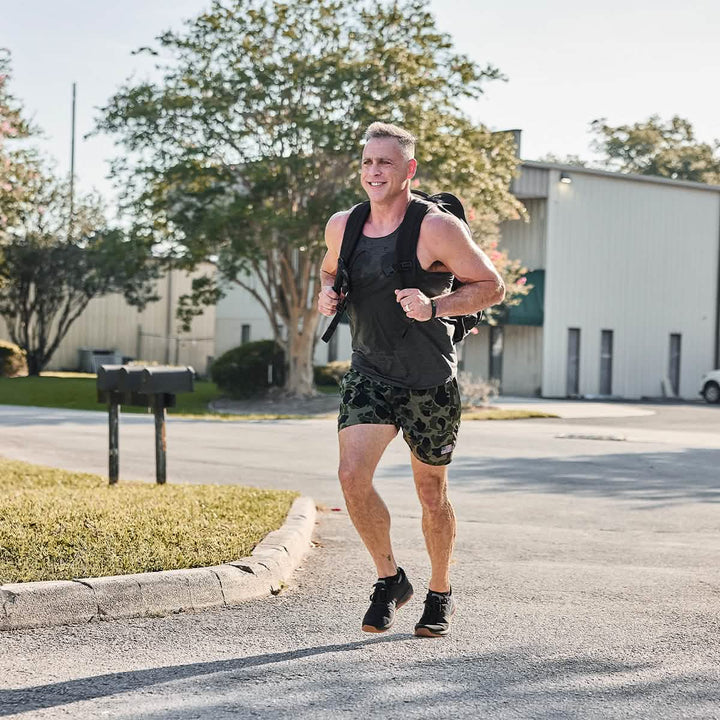 Under a sunny sky, someone jogs past buildings and trees, wearing the GORUCK Rucker 4.0, a black tank top, and camo shorts.