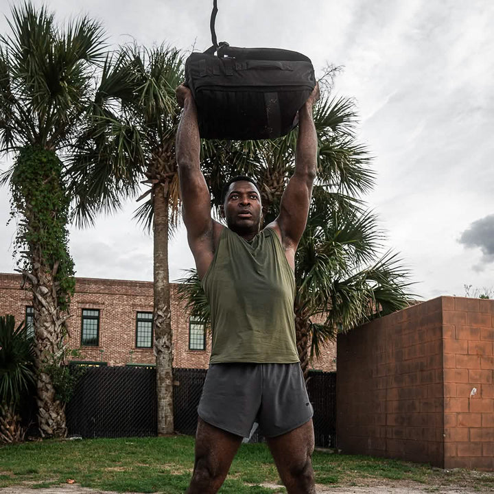 A man lifts a GORUCK Rucker 4.0 overhead outside, amidst swaying palm trees and classic brick buildings.