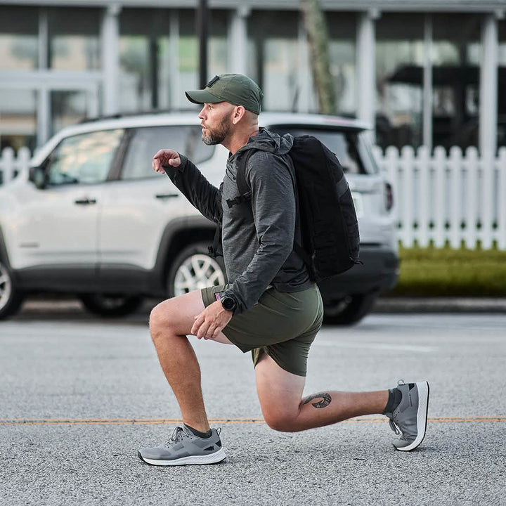 A man is lunging with a GORUCK Rucker 4.0 on a road, wearing a cap, gray top, and shorts in front of a parked SUV.