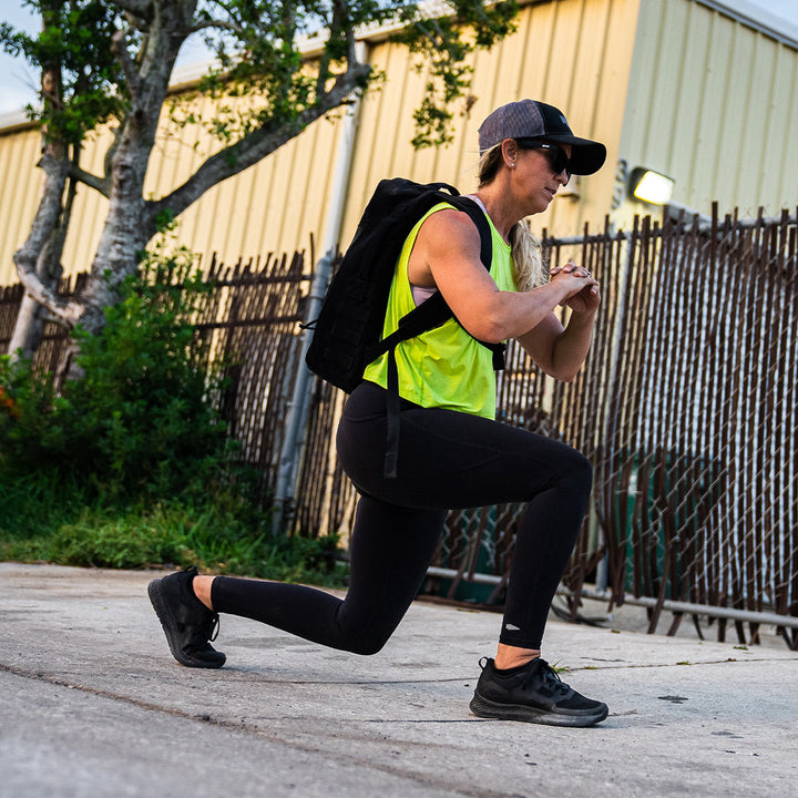 Dressed in activewear, a person is performing lunges outside near a fence, sporting a backpack and cap while their GORUCK Rough Runner - Blackout running shoes with a Gradient Density EVA midsole deliver comfort and support.