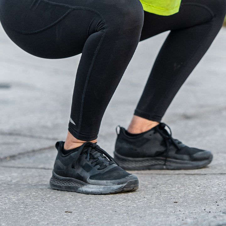 A person squats on a concrete surface, sporting black leggings and the sleek GORUCK Rough Runner - Blackout shoes, designed with a Gradient Density EVA midsole for enhanced comfort. The versatile road-to-trail outsole ensures they're ready for any adventure.